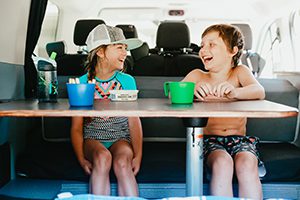 Two children enjoying a snack and beverage in the back of the vehicle with a dinette table attachment to place their wares.