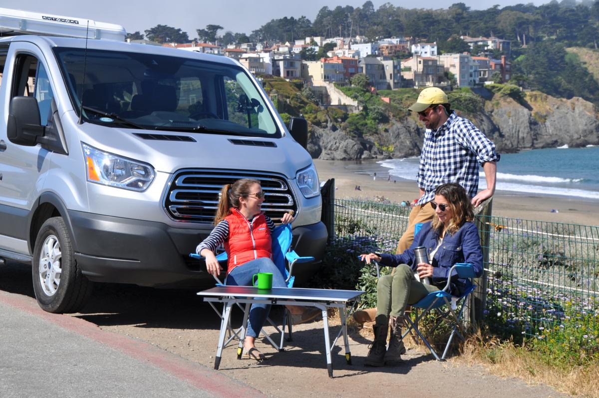 a group of people sitting around a camper van on the beach.