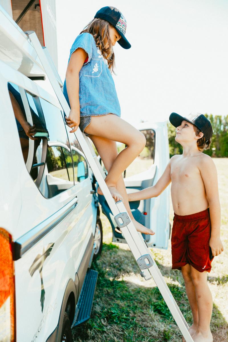 two kids standing on a ladder in front of a camper van.