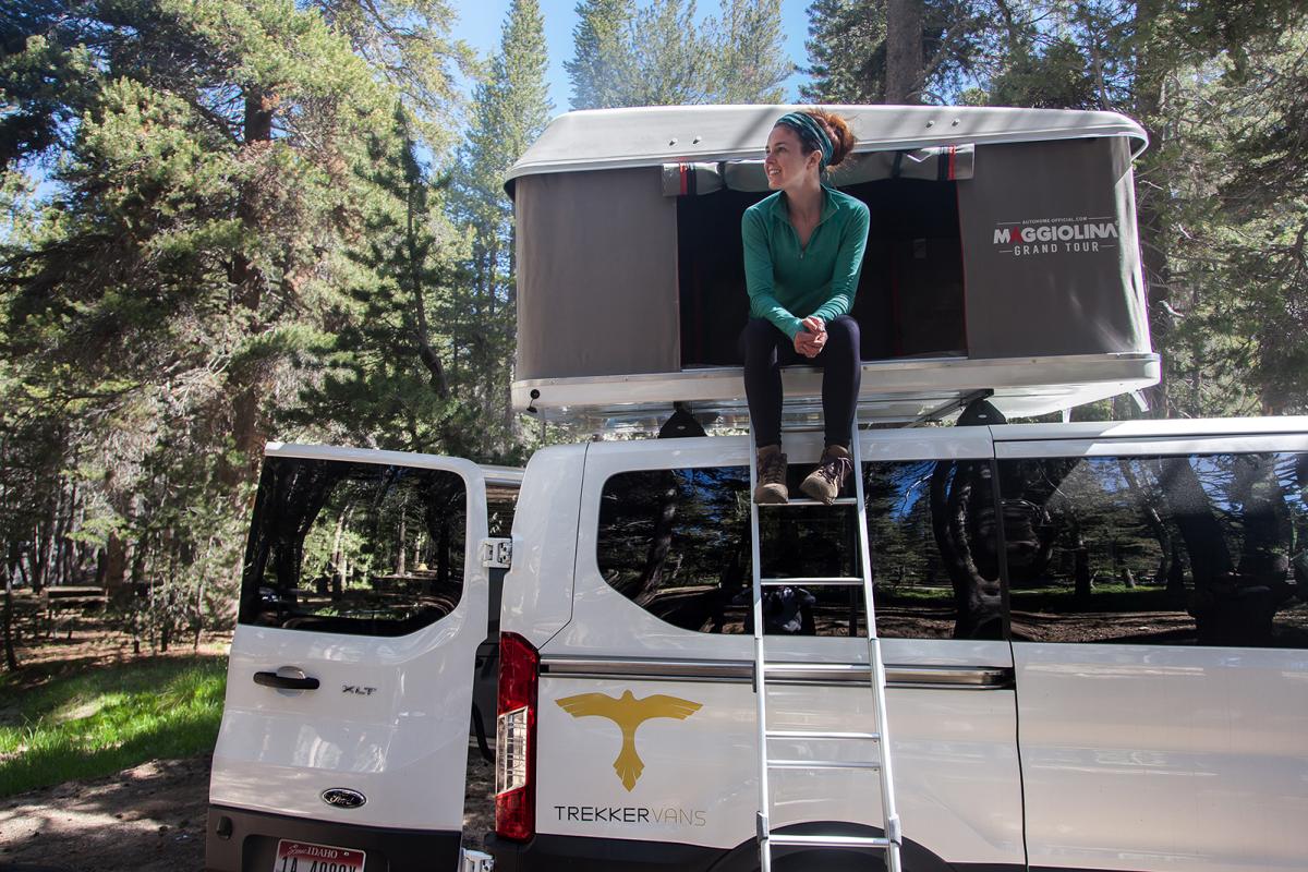 a woman sits on the top of a van with a tent on top.