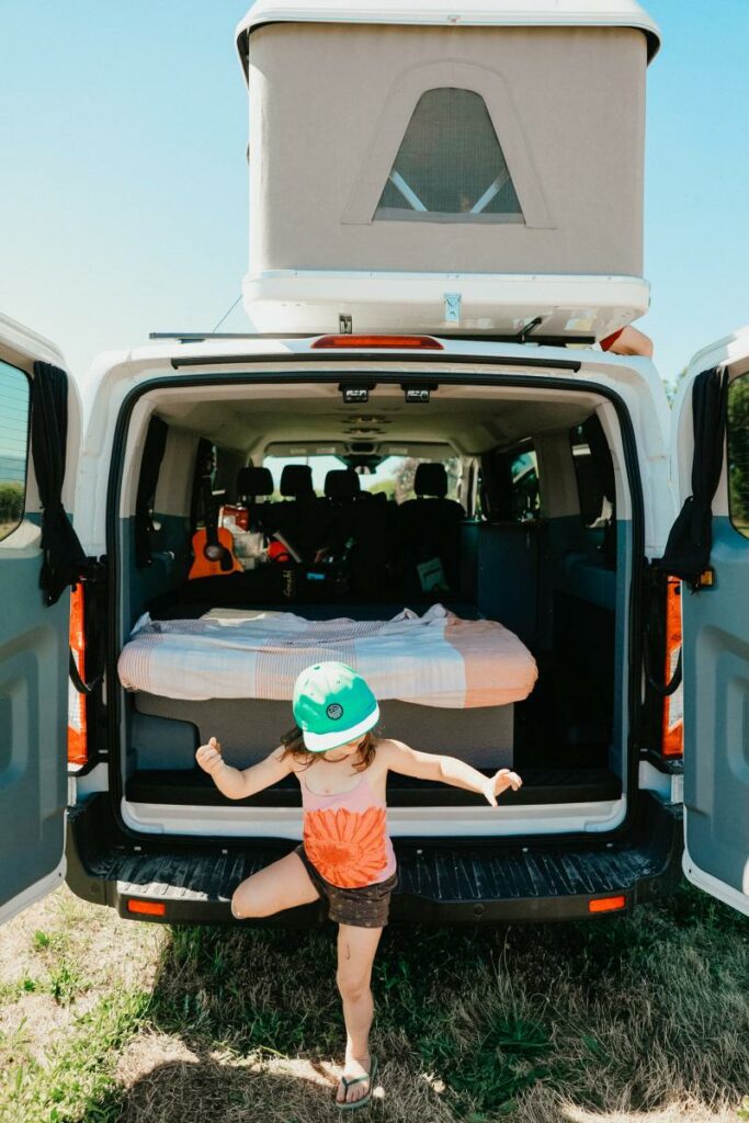 A little girl standing in the back of a van with a tent in it.